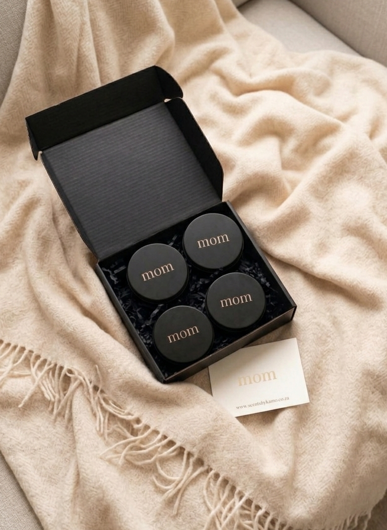 A luxury matte black gift box containing four 90g solid perfume tins with gold 'mom' branding, resting on eco-friendly shredded paper and a cream knit background.