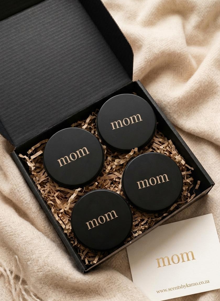 A luxury matte black gift box containing Four 90g solid perfume tins with gold 'mom' branding, resting on eco-friendly shredded paper and a cream knit background.